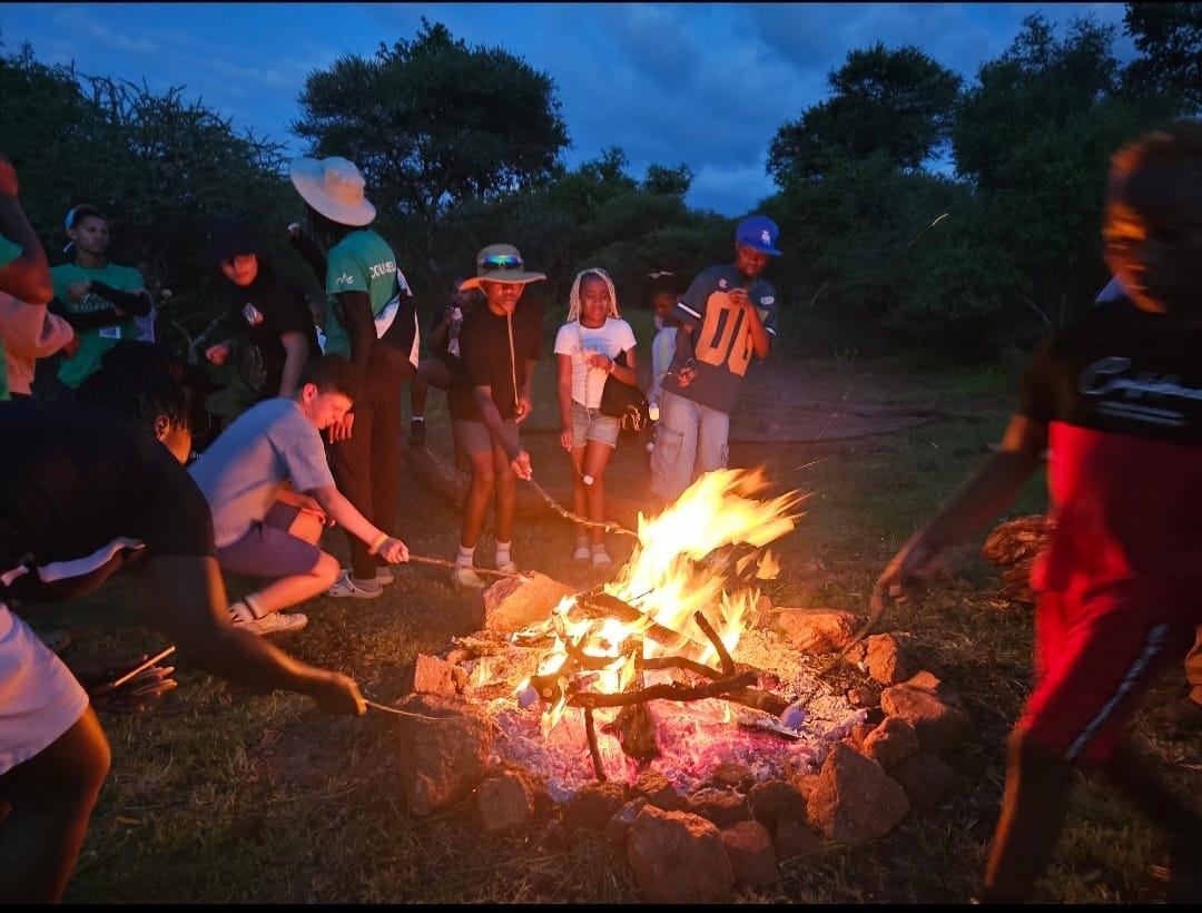 Campers holding long sticks over a rock-ringed campfire at dusk, bushveld trees behind