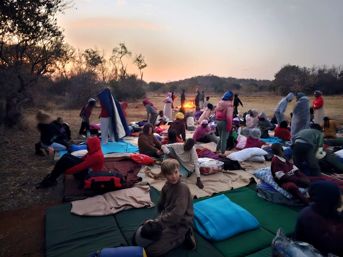 Campers in blankets on foam mattresses spread across open ground, a fire burning under an orange sky