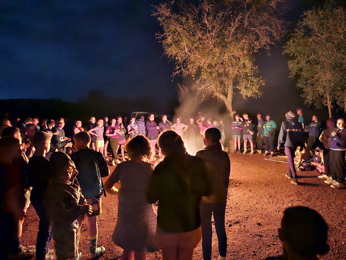 A wide circle of campers standing around a fire beneath a large tree at night