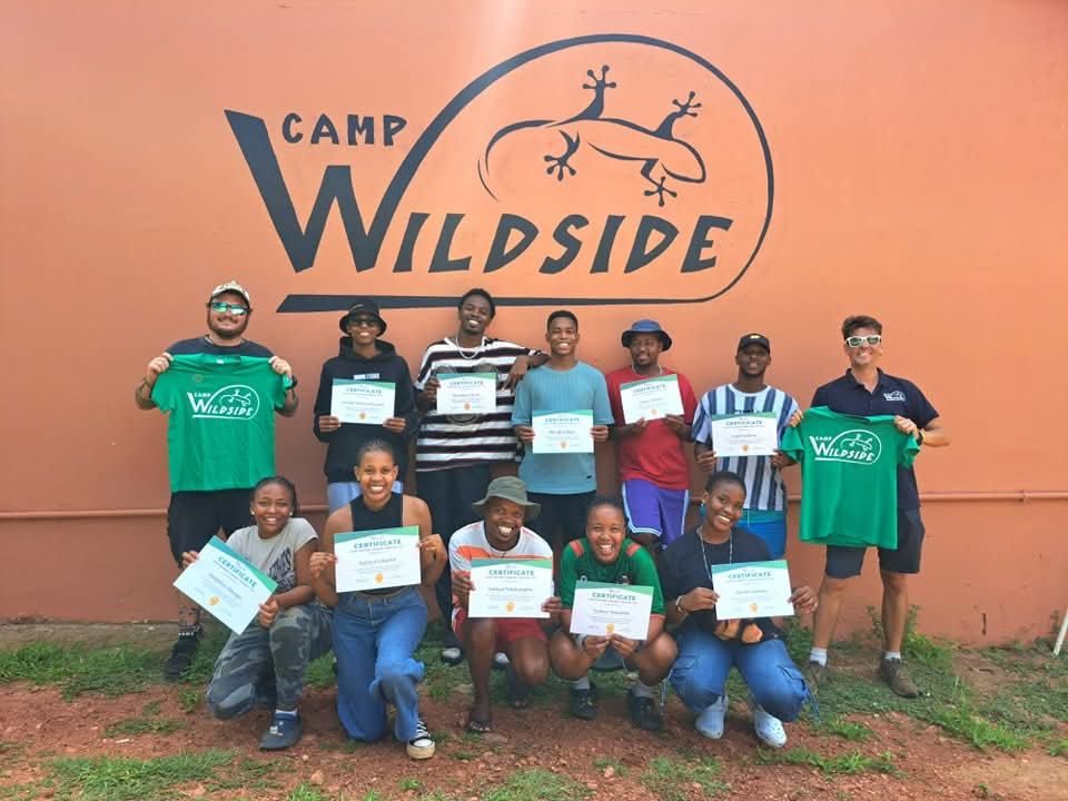 Twelve Camp Wildside counsellors in two rows holding their certificates in front of the painted camp logo wall