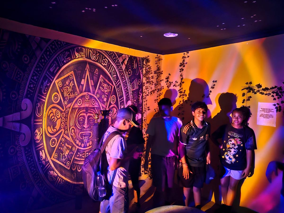 Children beside a large Aztec sun-stone mural under purple and orange lighting in the escape room