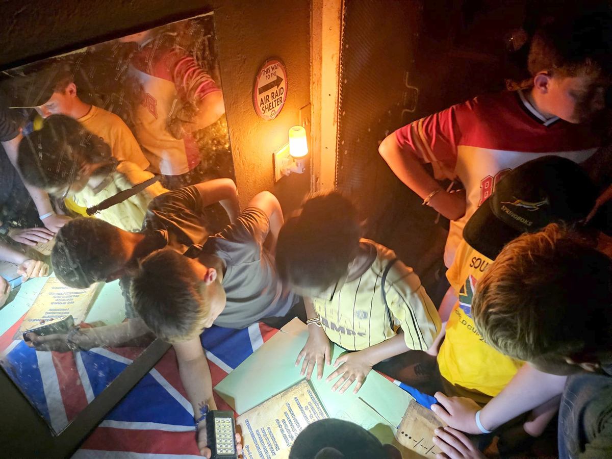Seen from above, children study clue sheets by torchlight beside an air-raid-shelter sign