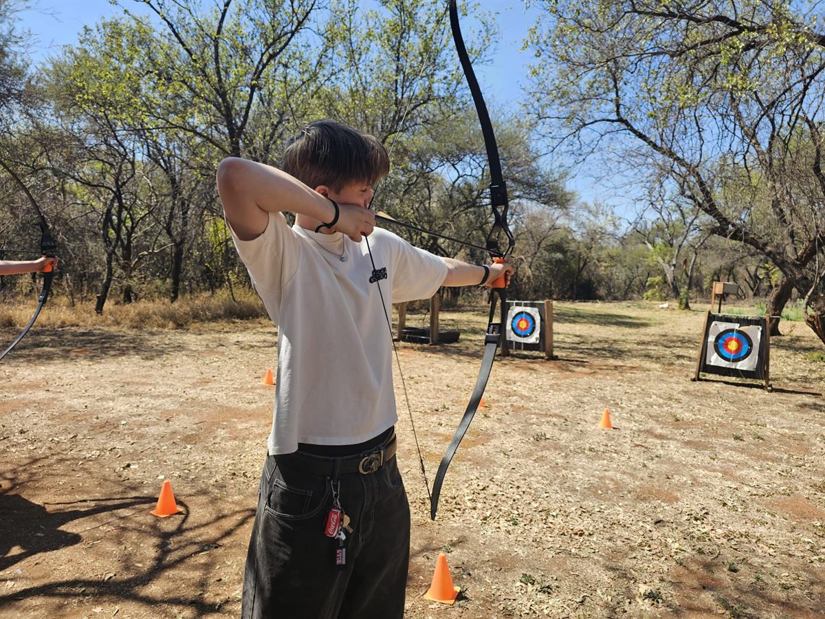 A teenager at full draw with a recurve bow on a dusty bush archery range