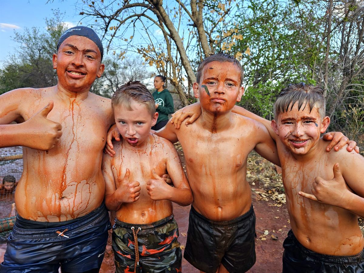 Four boys covered head to toe in red mud grin and give a thumbs-up after the obstacle course