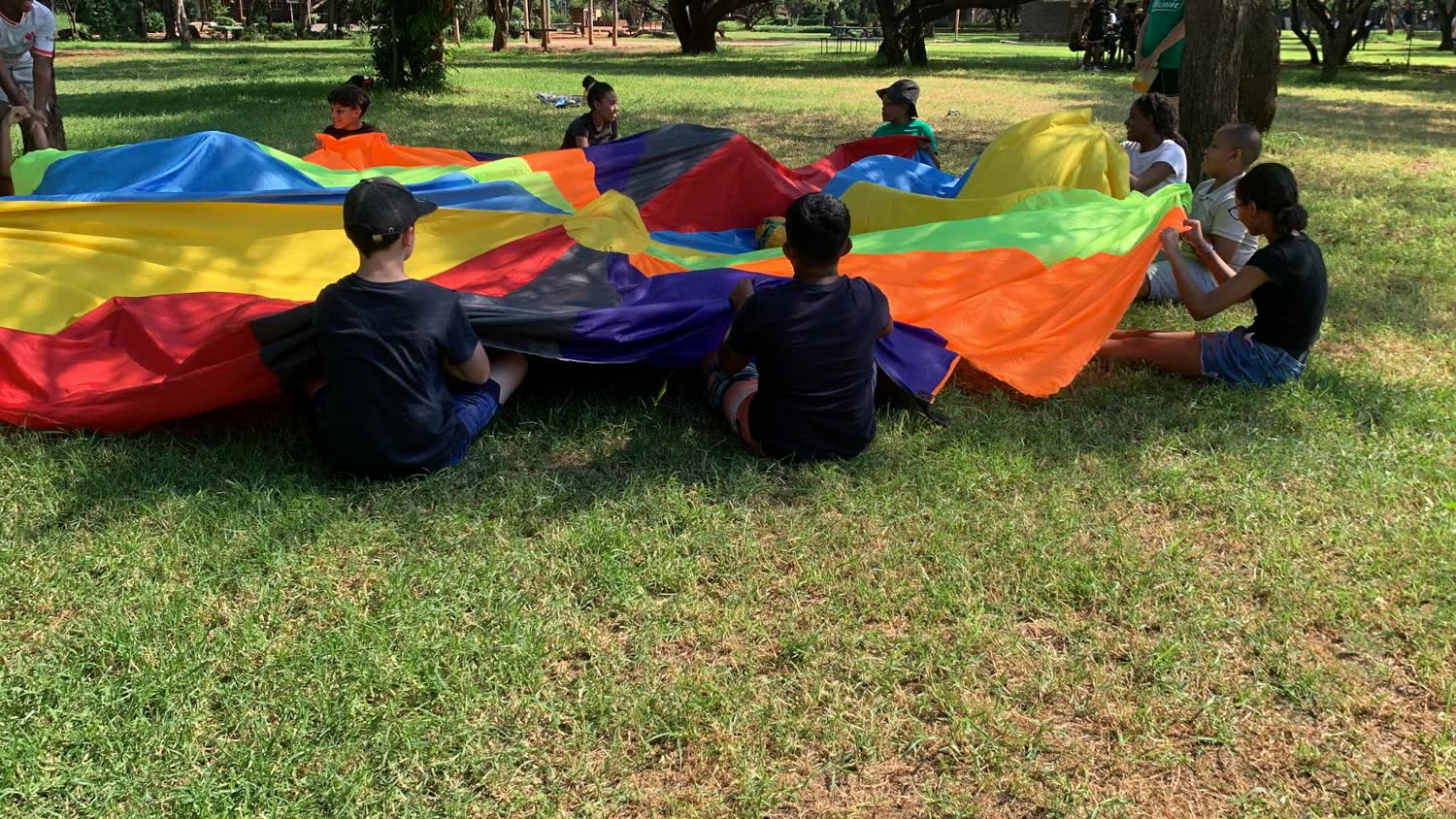 Campers sitting on the grass under a rainbow parachute during a group game