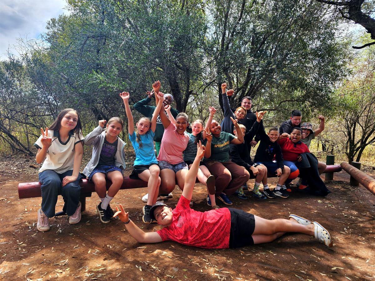 Campers sitting in a row on a wooden log beam with arms raised, trees behind