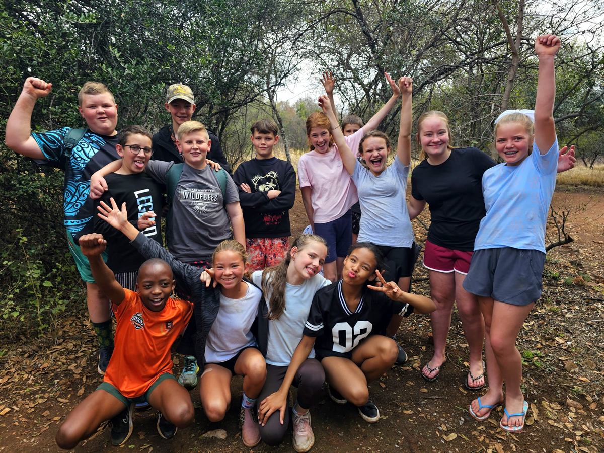 A group of campers posed on a sandy bush path, some crouching in front, others with arms raised