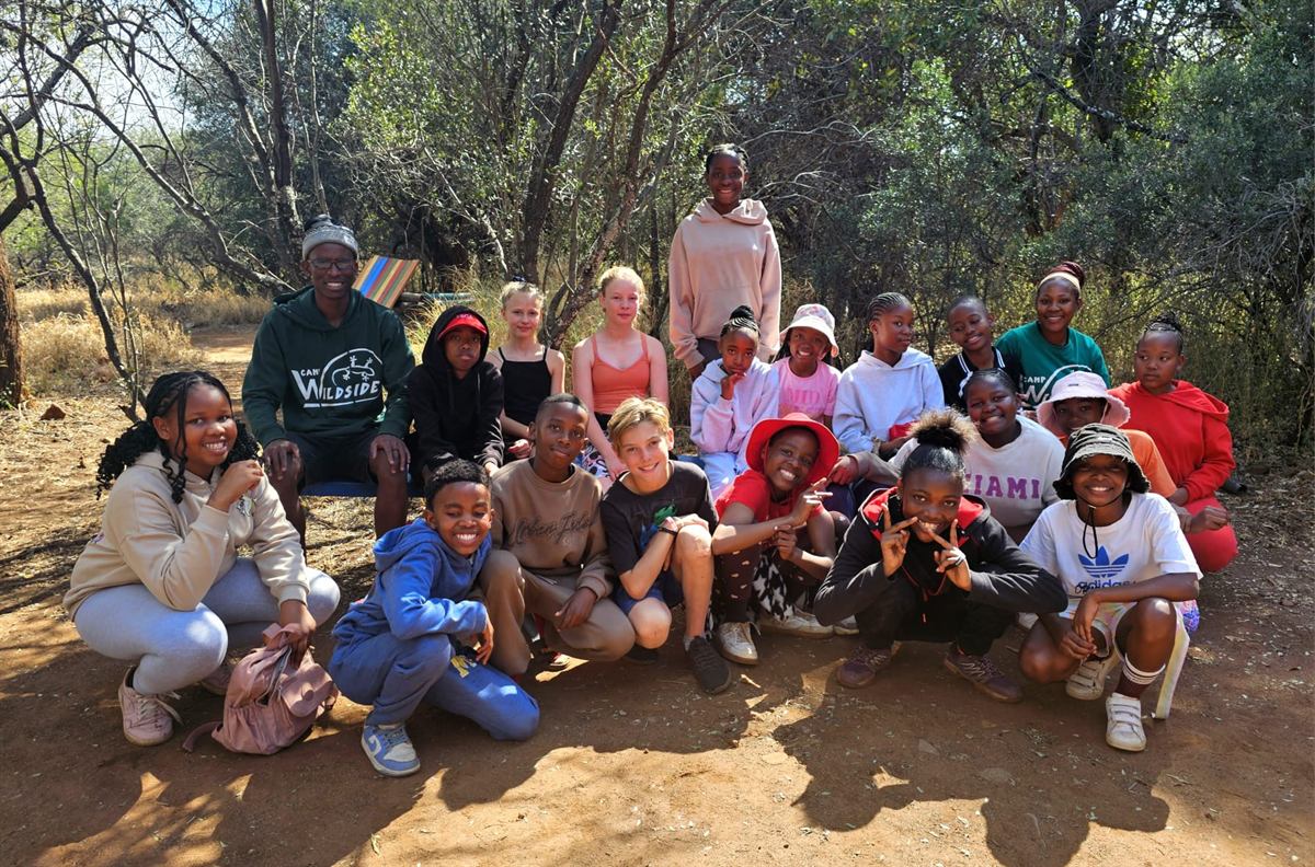 A large group of campers seated and standing in dappled shade among bush trees