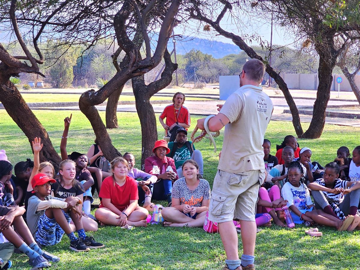 The reptile handler shows a small orange snake to campers seated on the grass under thorn trees