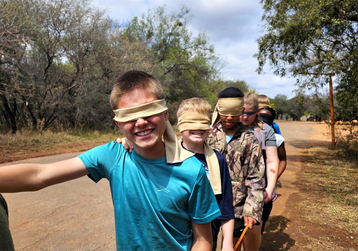 Blindfolded children walking in a line along a bush road, hands on the shoulders of the child in front