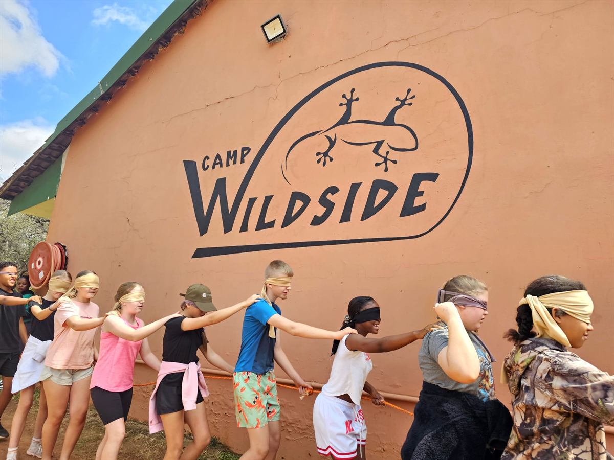 A line of blindfolded children walking hand-to-shoulder beneath the Camp Wildside gecko logo
