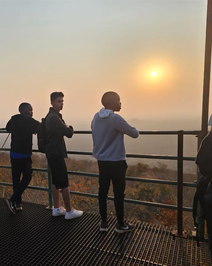 Teenagers at the railing of the hilltop lookout platform, the sun low over a hazy valley